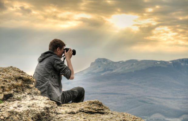 A photographer crouched on a rock, aiming a camera at distant mountains