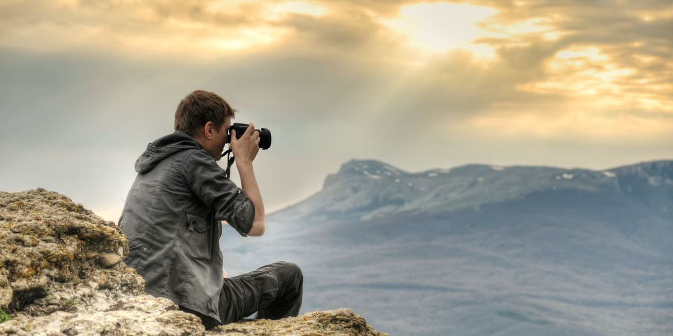 A photographer crouched on a rock, aiming a camera at distant mountains