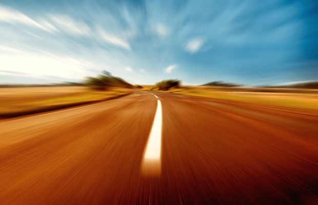 A straight road, blurred by speed, across flat orange ground under a blue sky